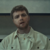 Alex Warren entrain de chanter, assis dans une laverie automatique dans le contexte du clip de sa chanson “Ordinary”.