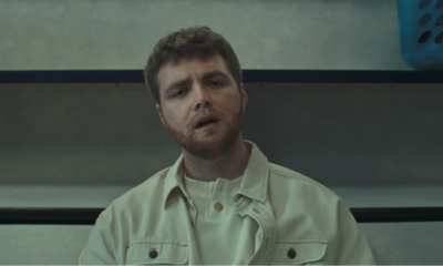 Alex Warren entrain de chanter, assis dans une laverie automatique dans le contexte du clip de sa chanson “Ordinary”.